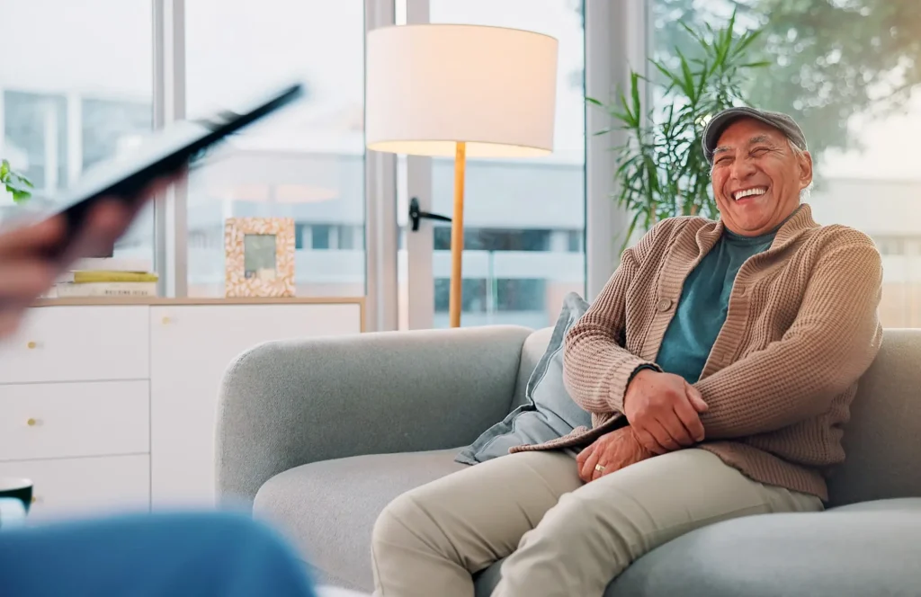 Older man in a cap laughing while sitting on a couch